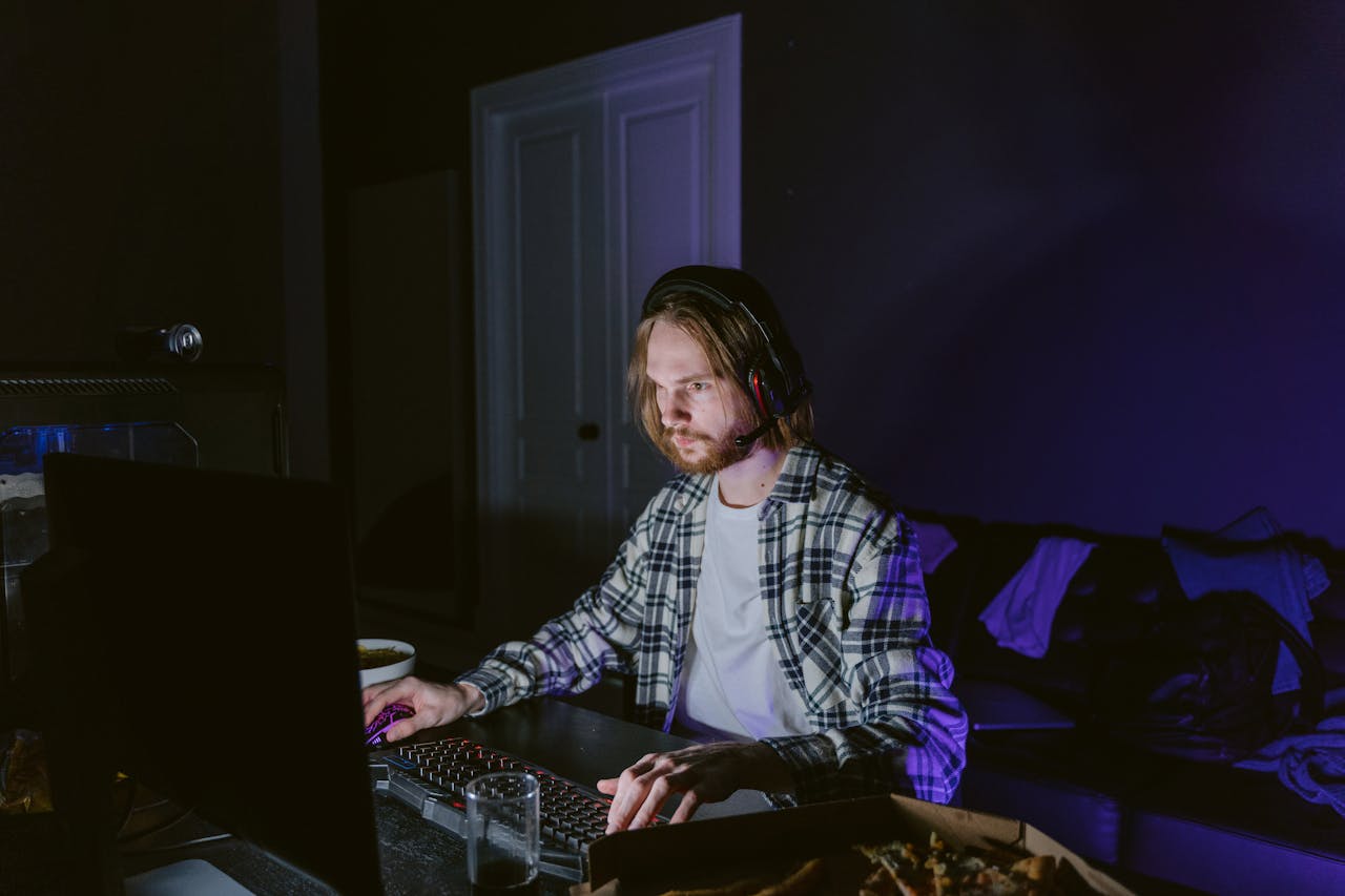 Features A man wearing headphones plays video games on a computer in a dark room, illuminated by screen light.
